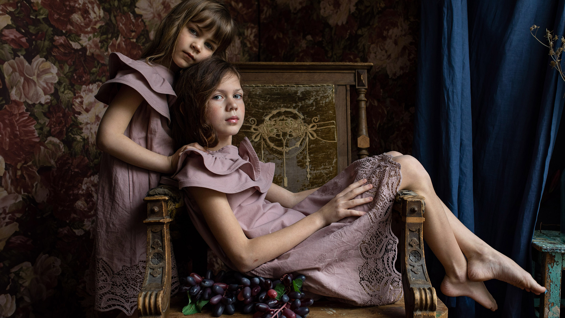 Two girls displayed in a room, featuring an old fashion theme, one girl sits on a chair and the other one stands behind her