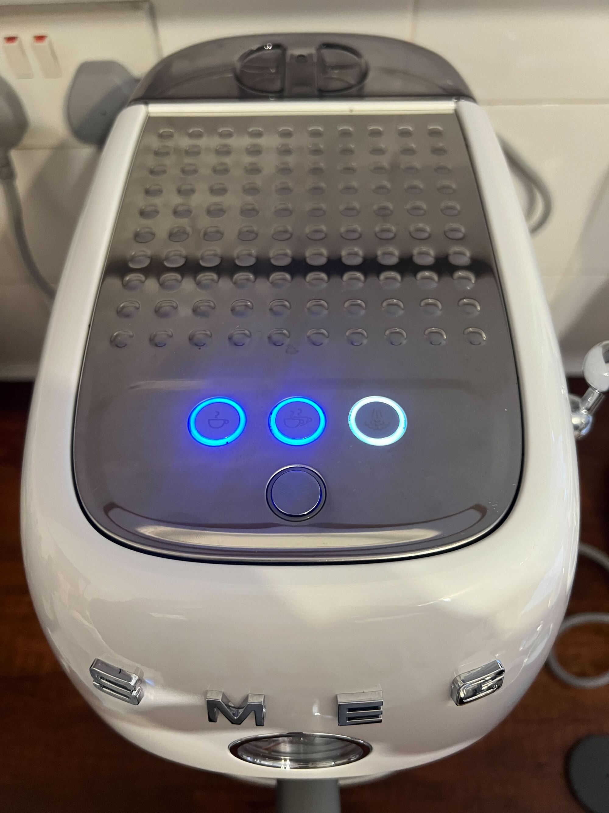 Close-up image of the top of a white Smeg cold brew coffee machine with the brewing buttons glowing in a blue light.