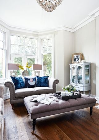 an elegant living room with a grey sofa, grey footstool, wooden flooring and white walls
