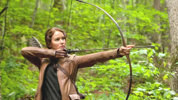 A still from the first Hunger Games movie in which Jennifer Lawrence plays Katniss Everdeen and is holding a bow and arrow in a forest.