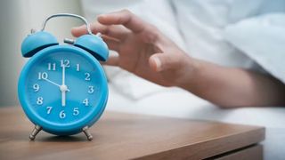 A person reaches over to a light blue alarm clock on a bedside table.