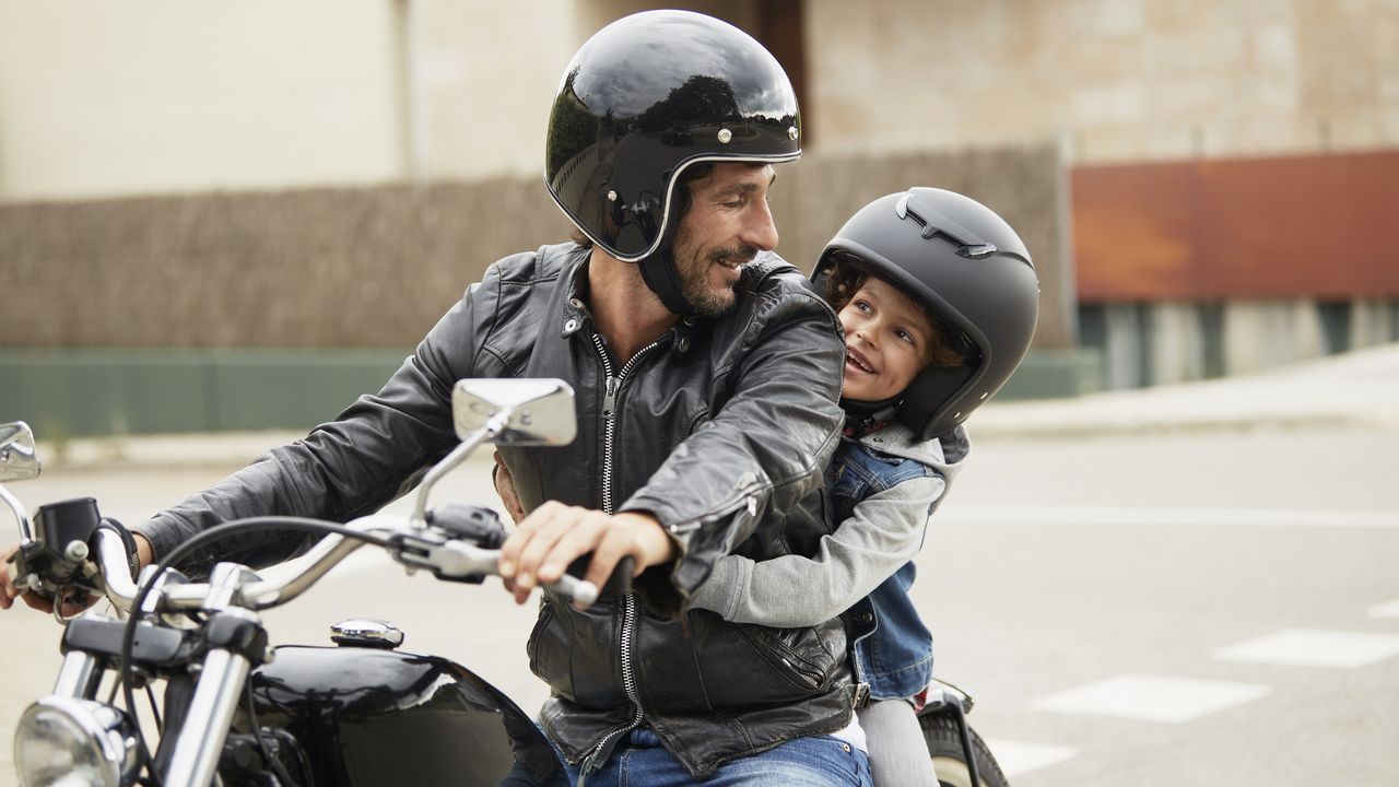 A helmet-wearing man on a parked motorcycle smiles at the child (also wearing a helmet) on the seat behind him.