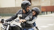 A helmet-wearing man on a parked motorcycle smiles at the child (also wearing a helmet) on the seat behind him.
