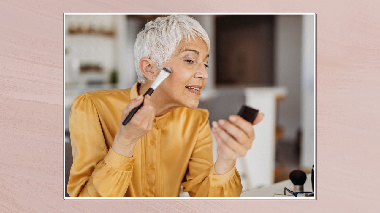 Side view of a woman with short grey hair applying makeup while looking into a small compact mirror