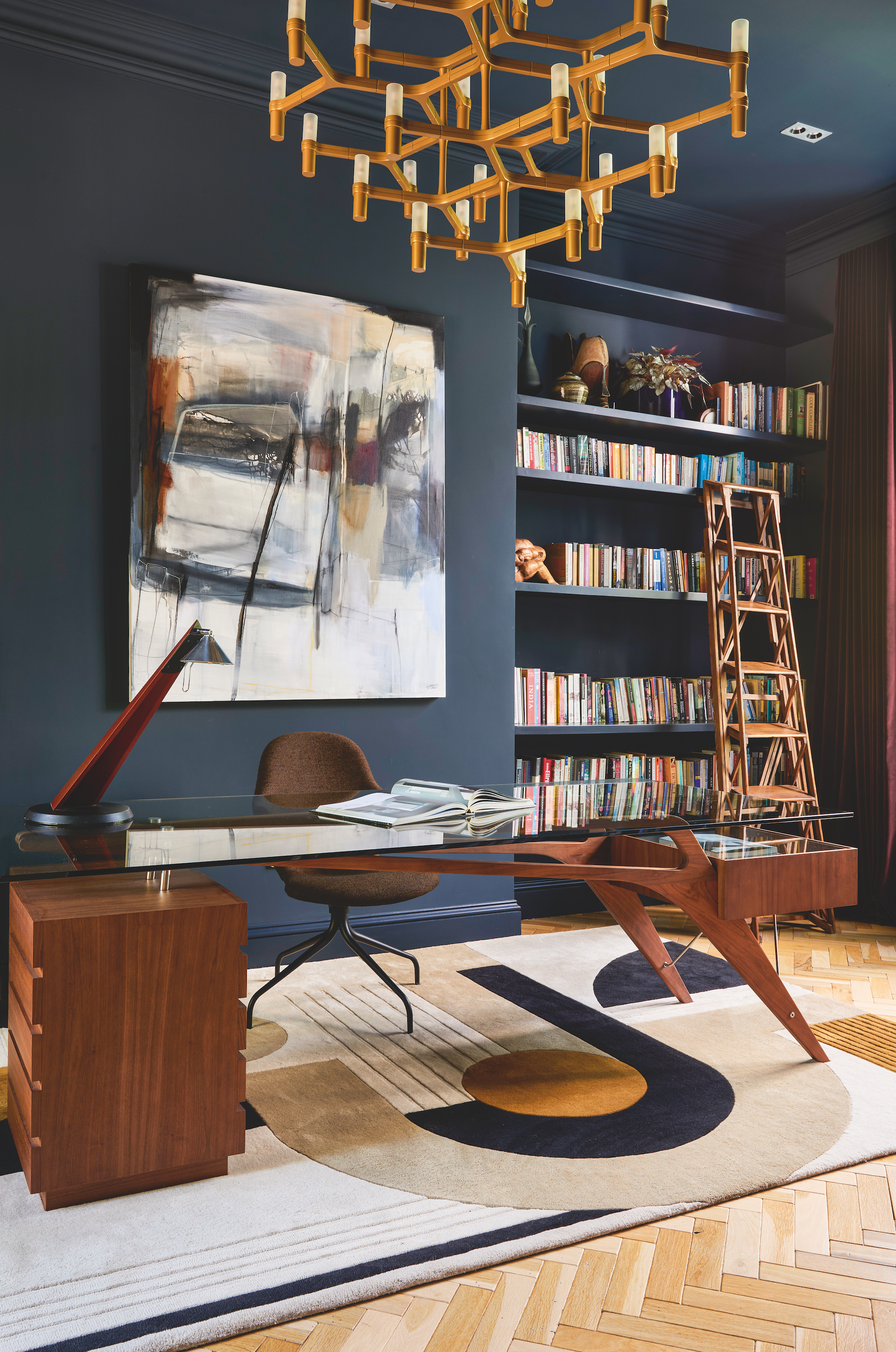 Navy, color drenched study with a bookcase in the right corner and a large glass and wood desk in the middle of the space.