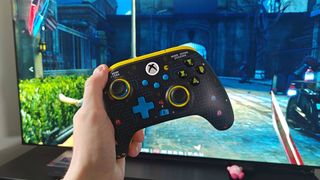 Hand holding Pac-Man PowerA wireless controller with TV in backdrop and Dishonored gameplay on screen.