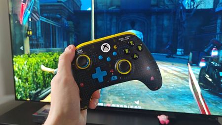 Hand holding Pac-Man PowerA wireless controller with TV in backdrop and Dishonored gameplay on screen.