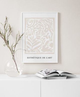 A wall art print with black writing on a beige wall above a white console with a book and a branch in a vase on it