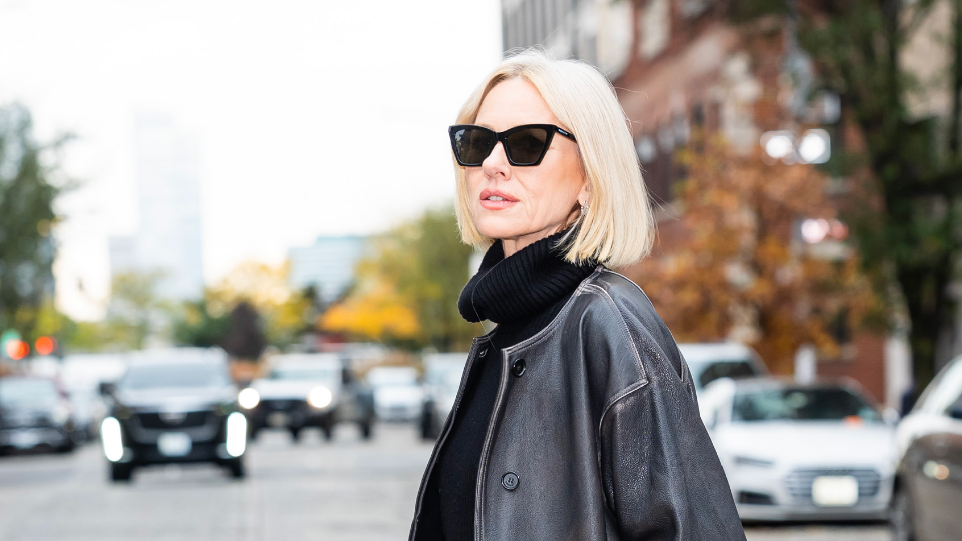 Naomi Watts wearing a turtle neck under a leather jacket and black cat eye sunglasses