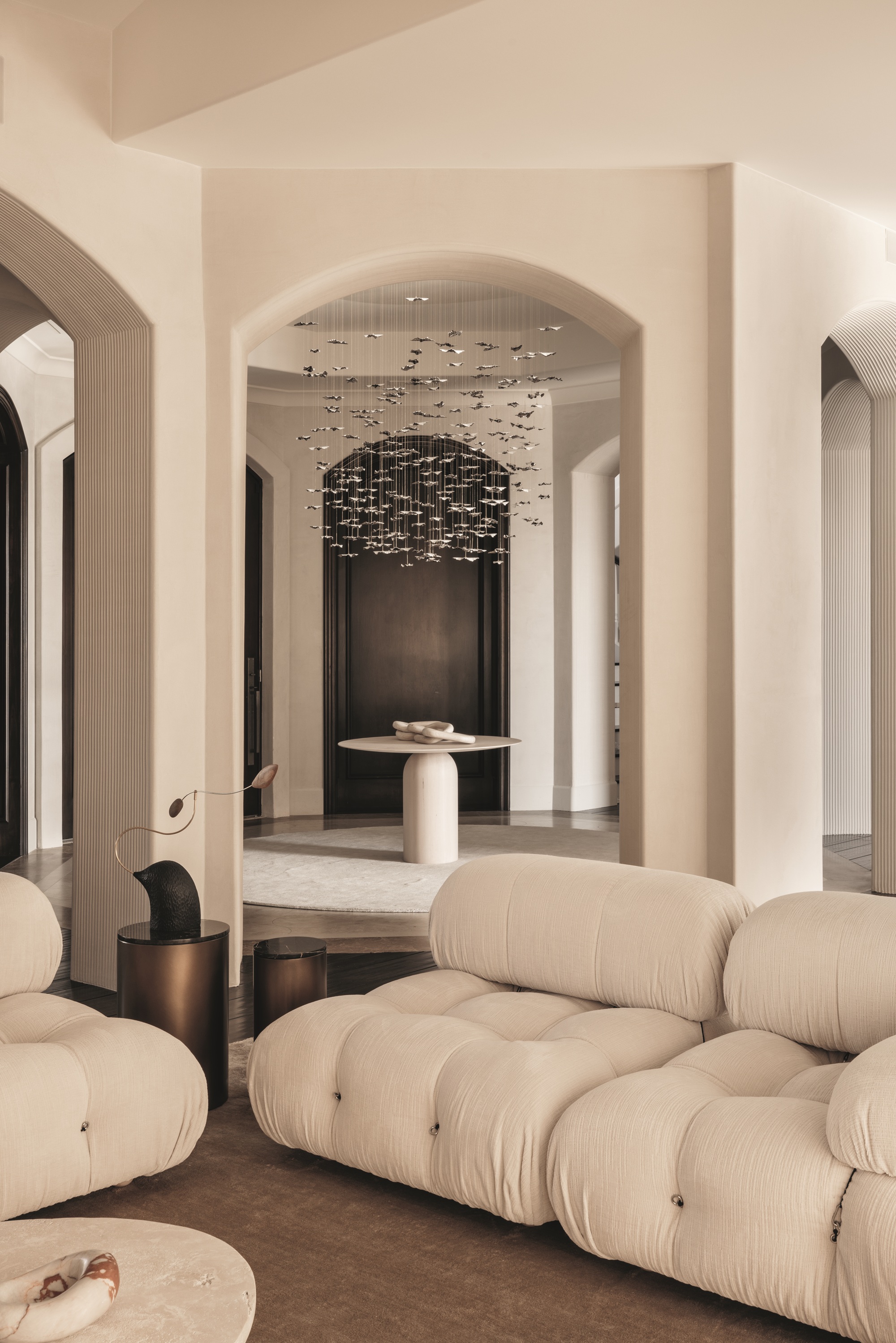 modern living room with beige modular sofa, bronze side tables, brown rug, and stone coffee table, with arched doorways leading through to the entryway with a round table, and sculptural hanging overhead