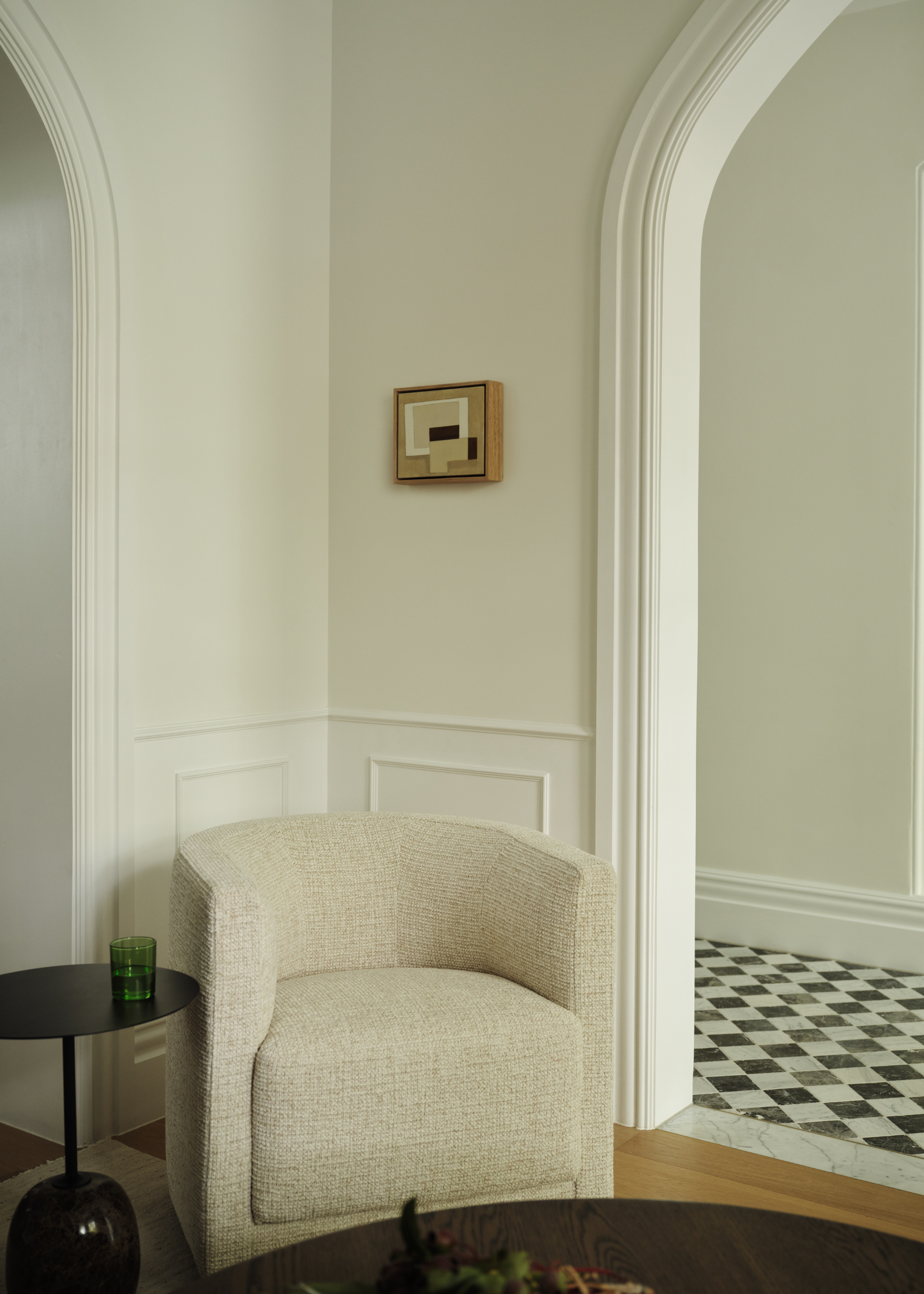 A white living room corner with a textured sofa chair, a black side table with a green glass, a wood coffee table, a mini framed art piece, and an arched doorway into a hallway with checkerboard marble flooring