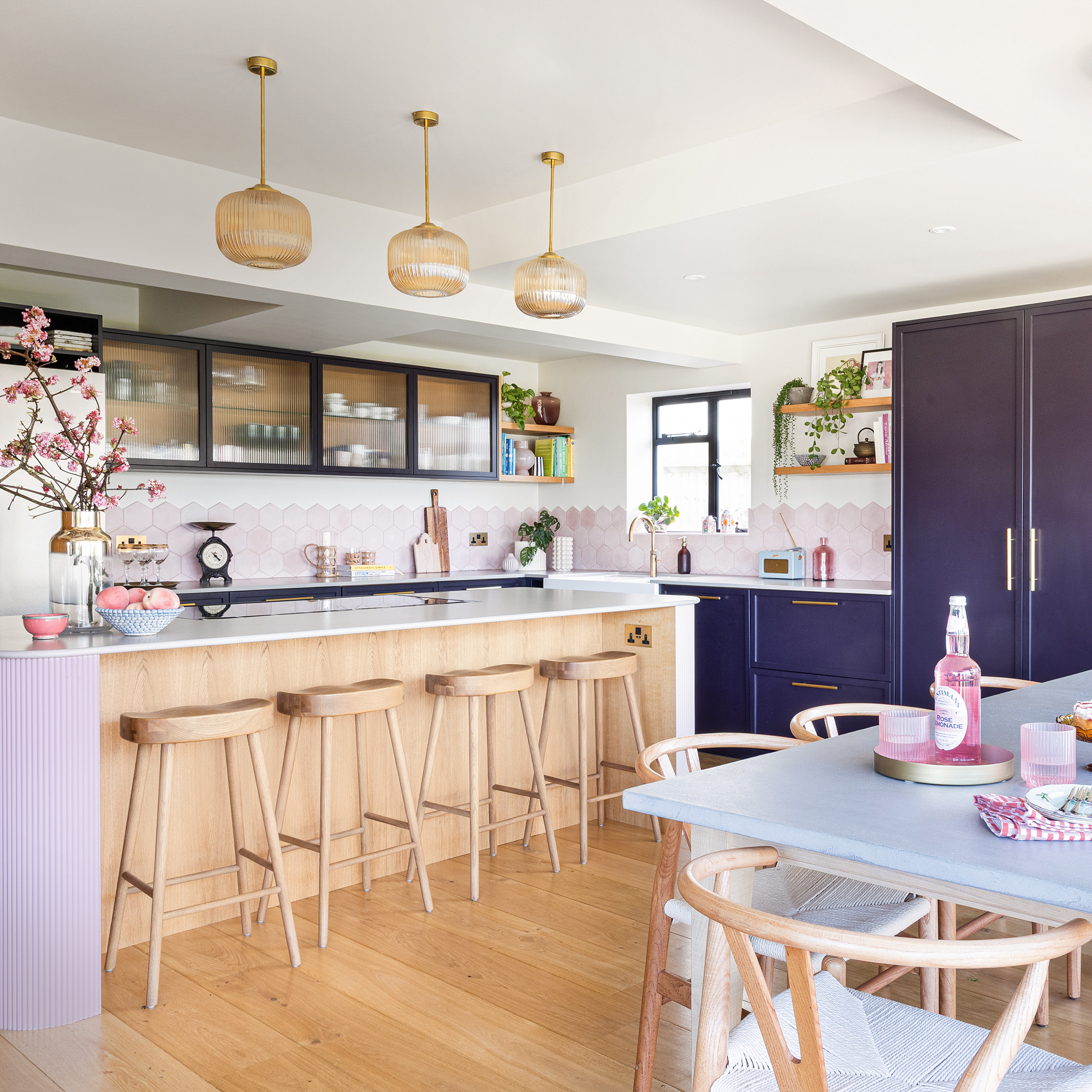 a purple and pink kitchen with purple cabinets and a pale pink hexagonal tiled splashback, pink island and a wooden dining. table and chairs