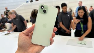 The back of a green iPhone 15 being held by the hand of TechRadar's Lance Ulanoff. It's held up in front of a blurred out crowd.