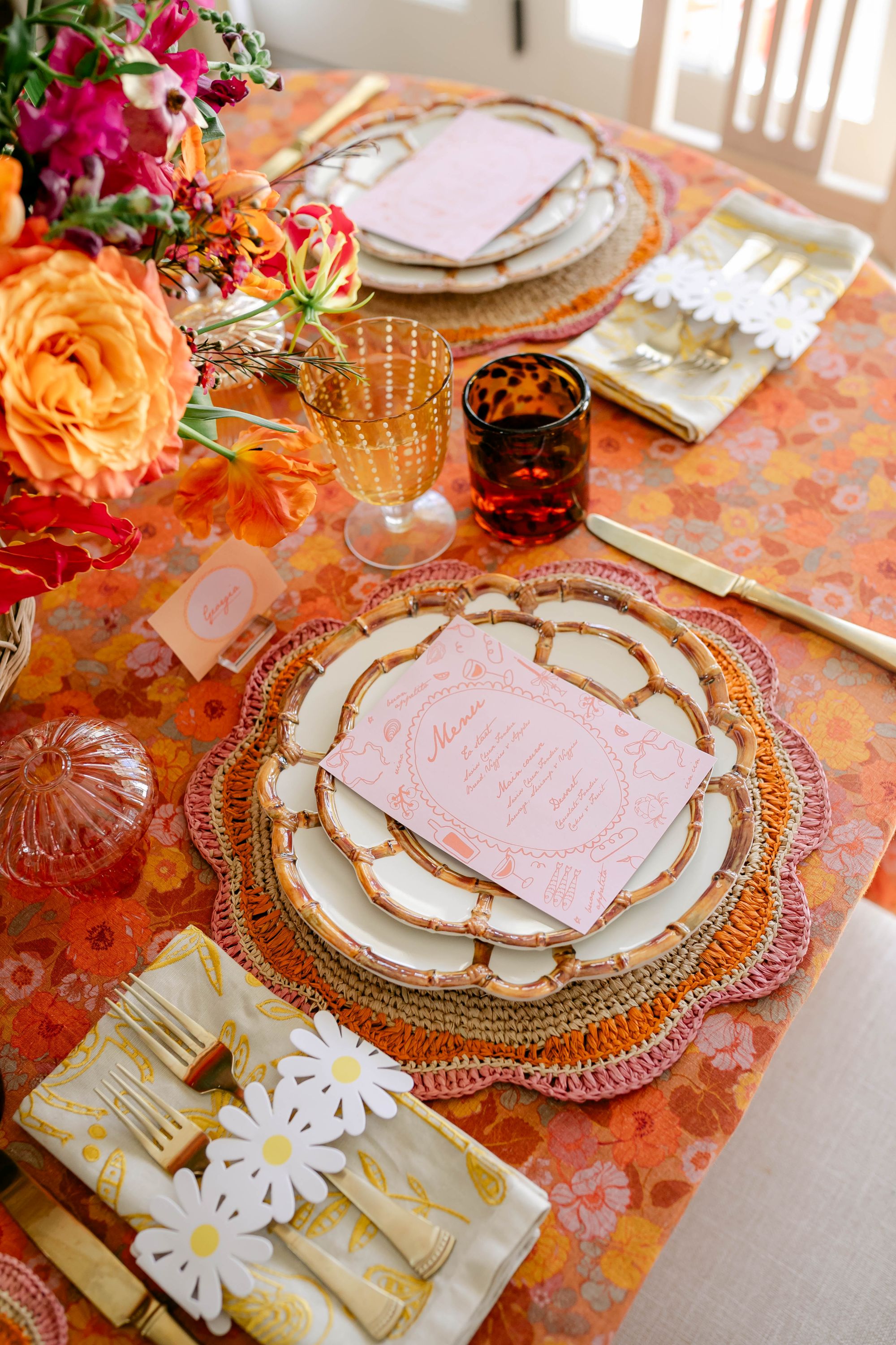 a tablescape with a floral orange table cloth, flowers, gold flatware, and white and brown plates