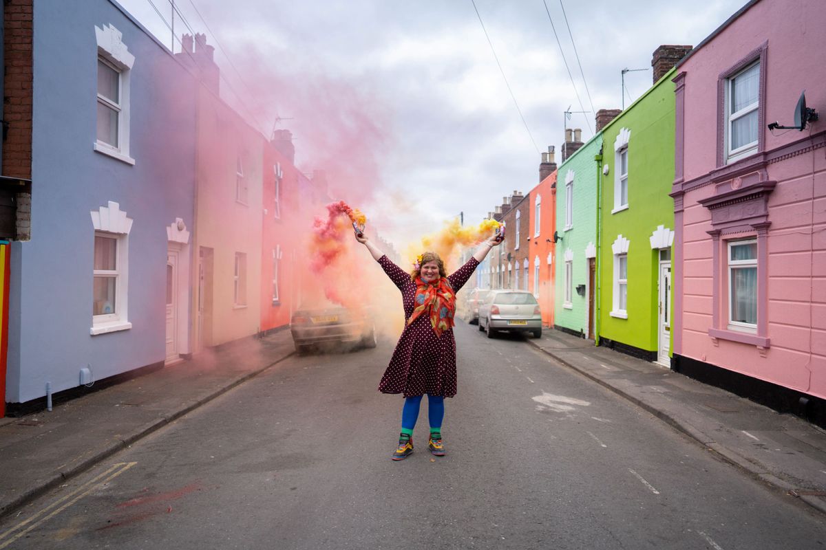 Woman in street with paint flare standing in between houses either side painted an array of colours