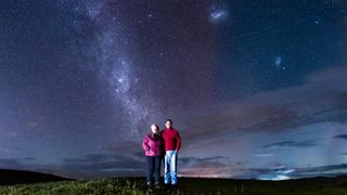 two people stand smiling at the camera against the background of the milky way stretching into the sky and a distinct red glow behind them, a diffuse glow filling the sky.