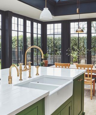 A light-filled kitchen with green cabinetry, a white Belfast sink, and brass taps. A large island overlooks a dining area, framed by floor-to-ceiling black windows.