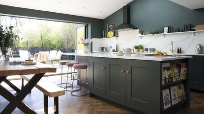 Open-plan kitchen diner with dark grey-green Shaker kitchen, parquet flooring, industrial-style dining table and benches, pink bar stools and bifold doors out to a decked seating area