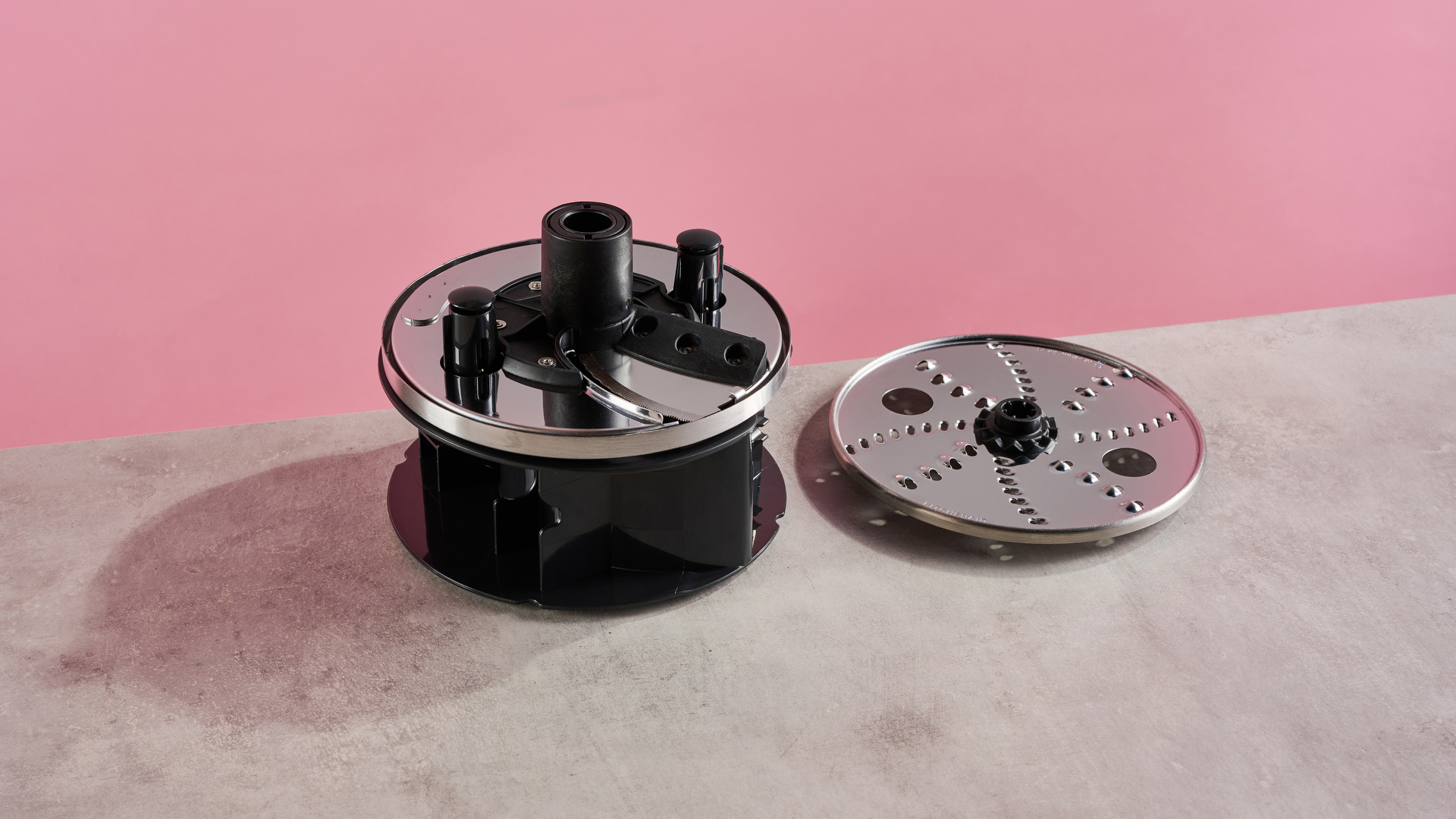 Image shows the black plastic storage caddy from the KitchenAid 13 cup food processor. It has the adjustable slicing disc on the top, and the reversible grating disc on the grey stone-effect surface on the right. It is photographed against a pink background.