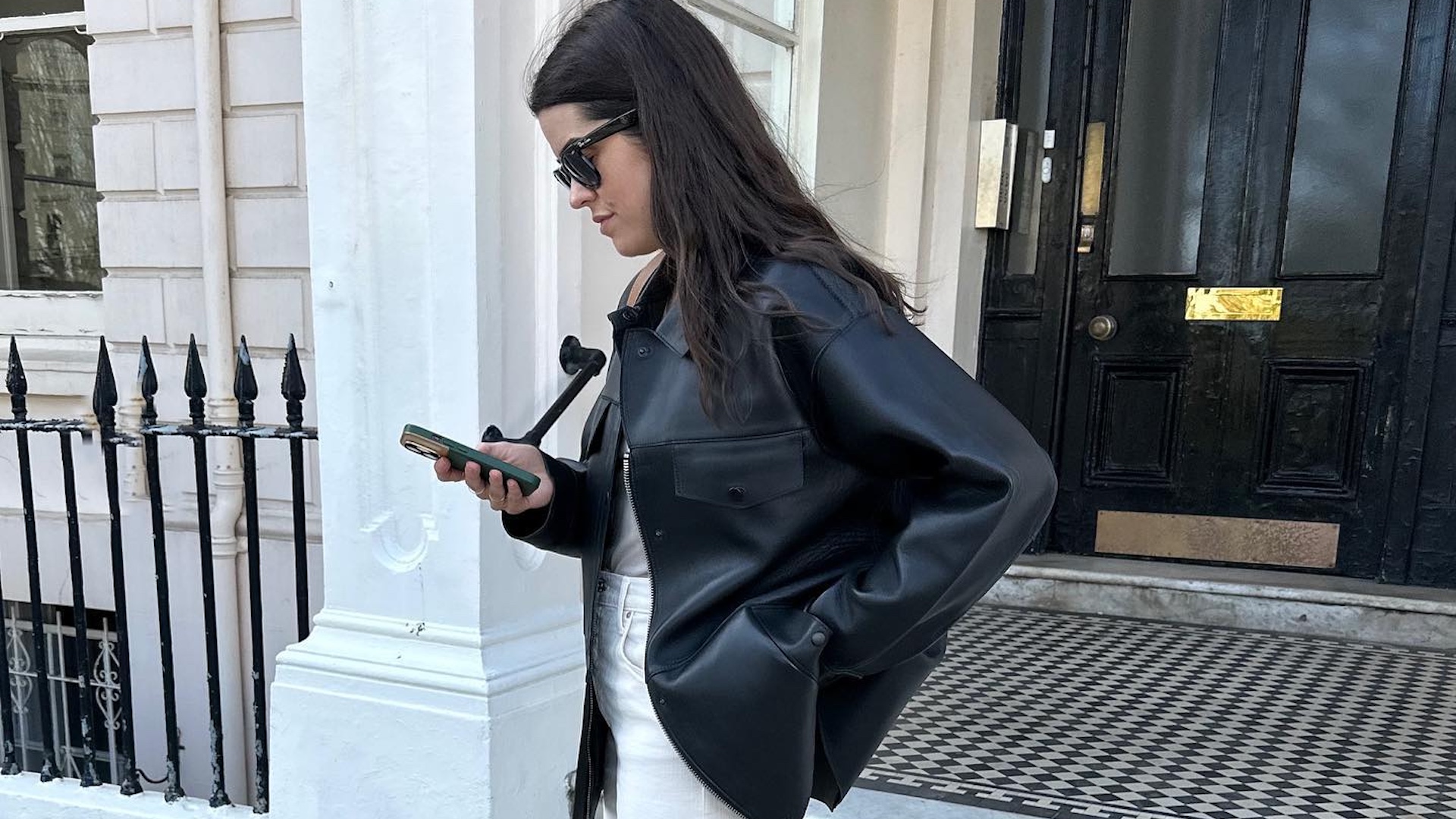 British style influencer Anna Howard poses on a London street looking at her phone wearing a black leather jacket and white jeans.