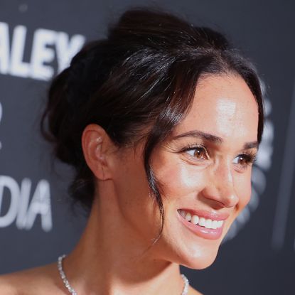 A headshot of Meghan Markle wearing a diamond necklace and a curly updo in front of a black Paley Center for Media background