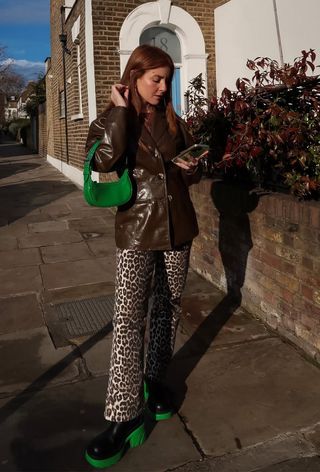 how to wear chelsea boots is shown in a photo of a woman standing on the sidewalk wearing a brown leather blazer with a bright green bag, leopard print trousers, and Chelsea boots with chunky green lug soles