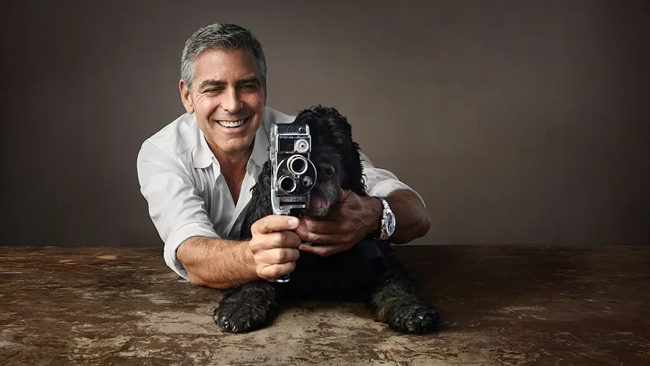 A man smiling and holding an old-fashioned film camera with a black Cocker spaniel resting its head beside him.
