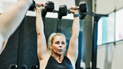 A woman stands holding two dumbbells overhead with straight arms.