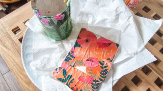 A decoupage green floral tin and two coral colored coasters on top of a white circular plate with kitchen towel, with a wooden grid table beneath them