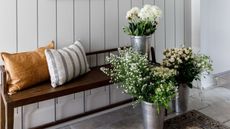 Small entryway with wooden paneled walls painted off white, with wooden bench and metal vases of white flowers