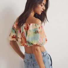 A model looking back over her shoulder in the direction of the camera wearing a colourful off-the-shoulder crop top with high-waisted denim jeans from New Look