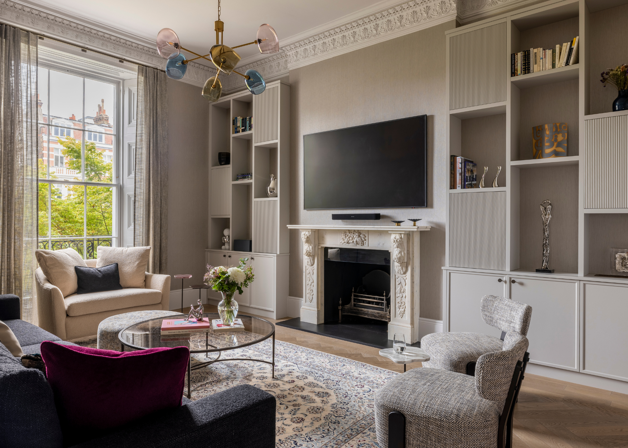 A modern living room with neutral walls and cabinetry with a dark sofa and a round glass coffee table with books and a clear glass of fresh flowers