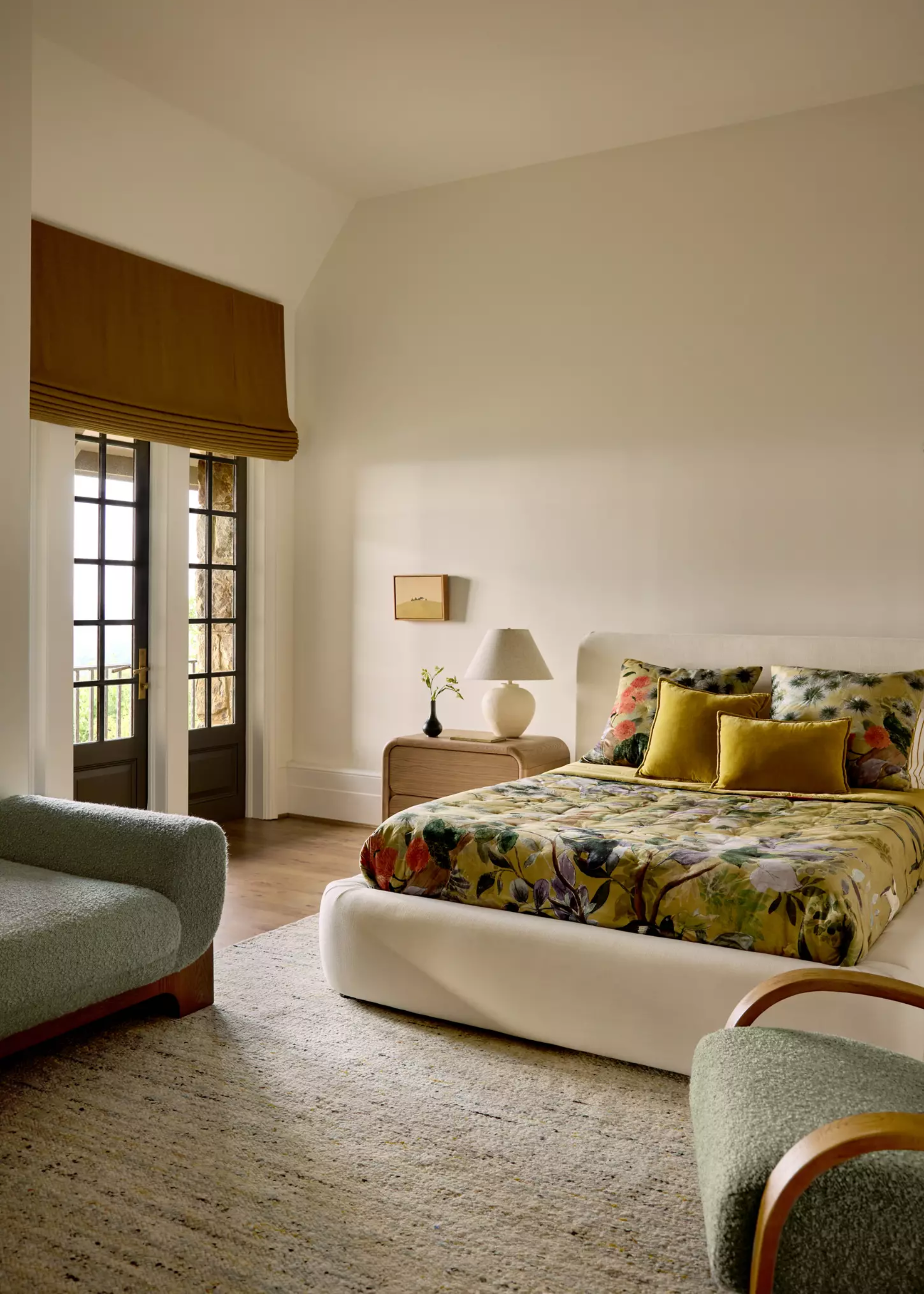 A white bedroom with balcony doors, a white bedstand, a floral bedscape, a wood side table with a white lamp and a vase, a sofa chair and an upholstered bench