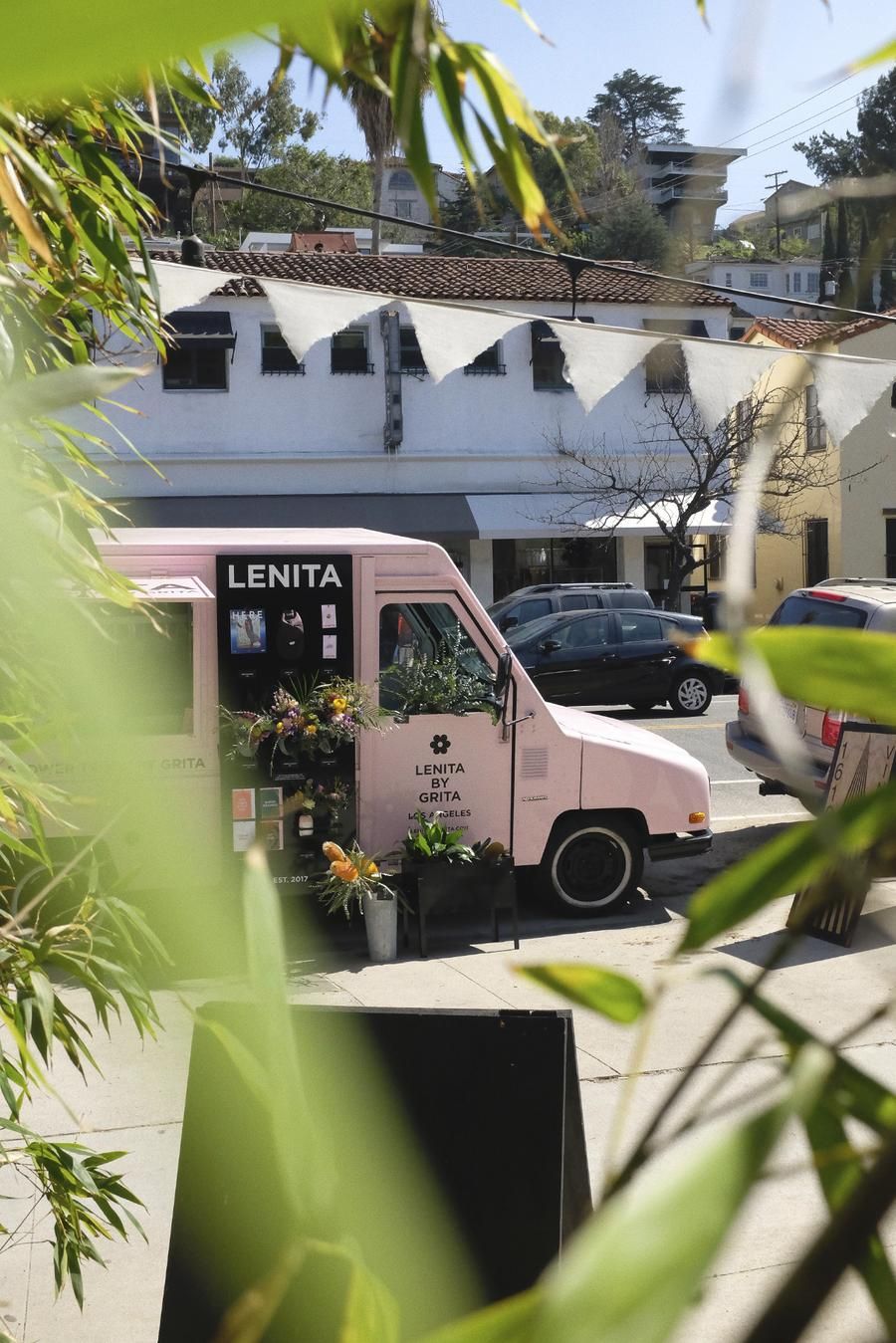 A flower shop truck in dust rose sits in the sunshine surrounded by plants.