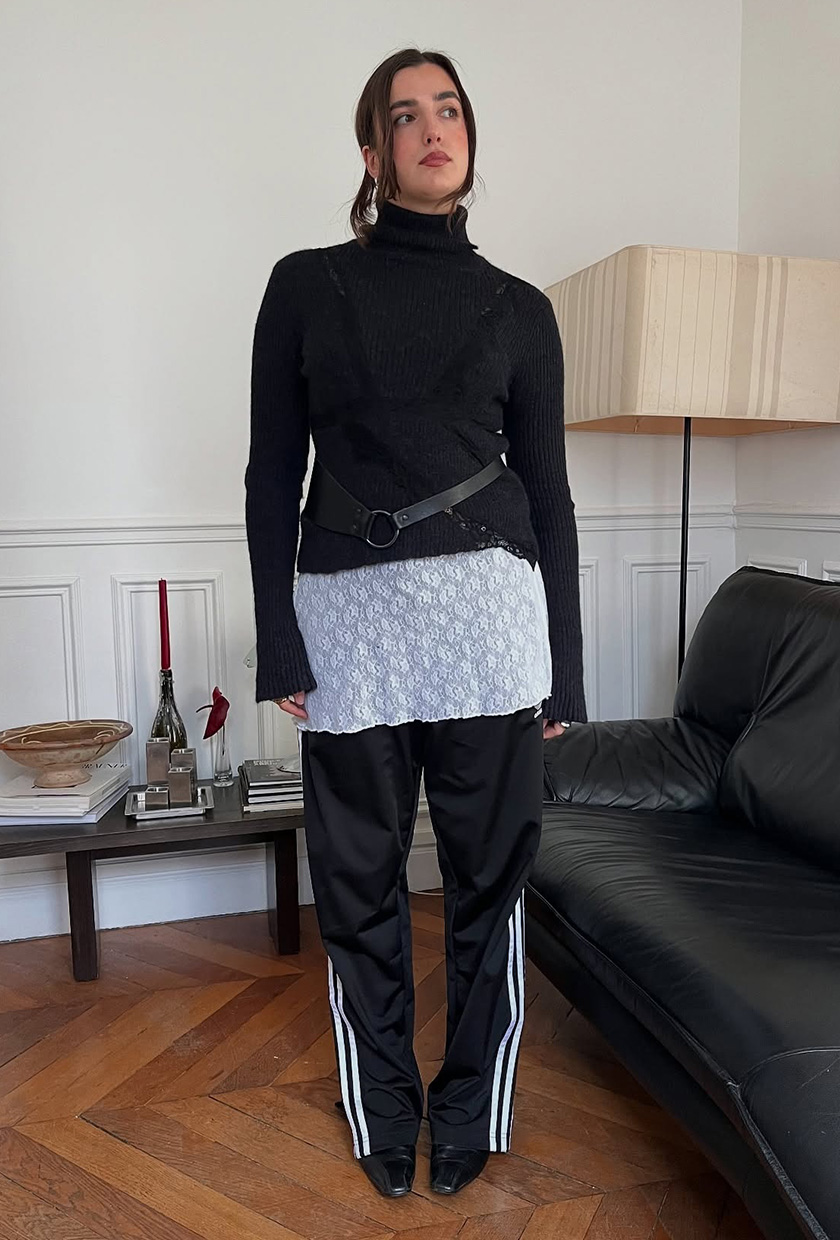 A woman standing inside her living room by a black leather couch wearing a black turtleneck sweater layered on top of a long sheer lace minidress and black track pants styled with black pointed ankle boots and a black belt.