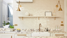 An off white kitchen with gloss metro wall tiles above a marble worktop and brass wall lights either side of an open shelf 