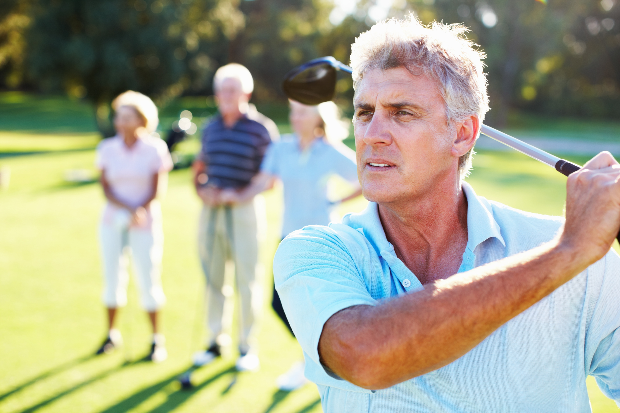 Handsome, mature man swinging his golf club with friends in blurred background