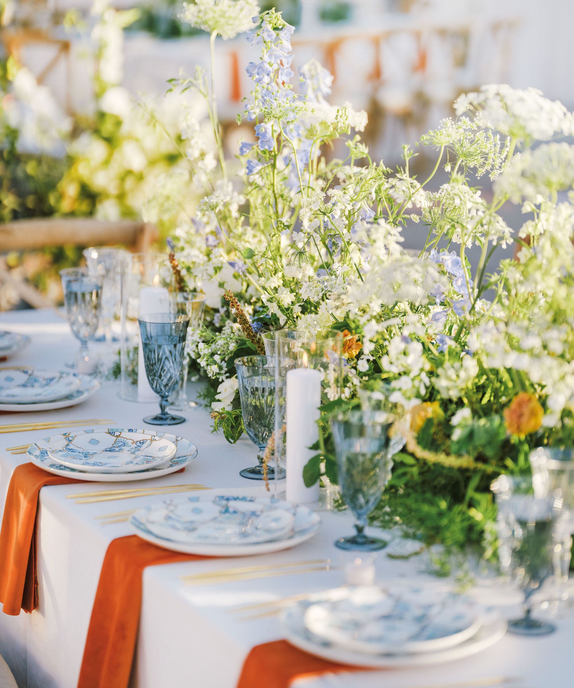 A long table with a white tablecloth, blue and white china plates, orange napkins, blue glassware and a large floral arrangement at the centre with dainty white, purple and orange flowers.