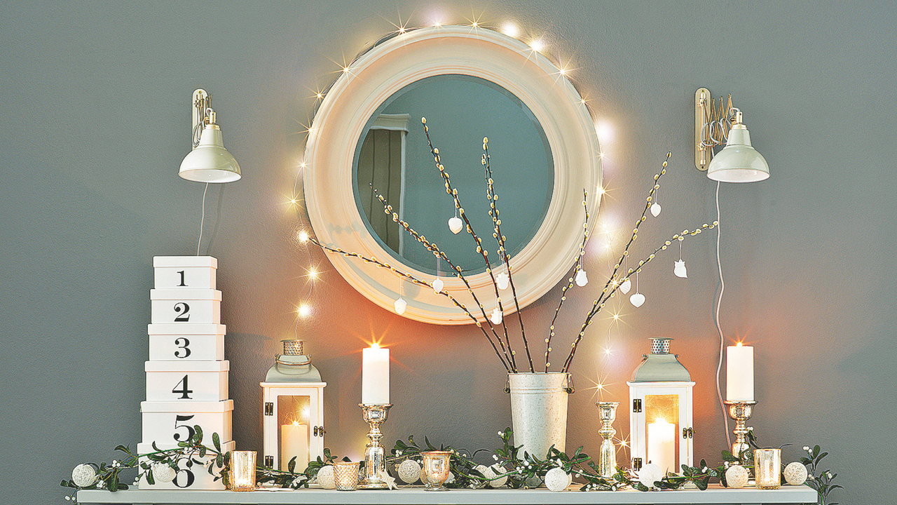 Dark grey console table display, garden utensils, large circular mirror with fairy lights, plant Christmas decoration, wall mouted lights, candle lanterns.