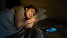 person sleeping in bed on their side facing the camera, with a phone on the bedside table in front of them