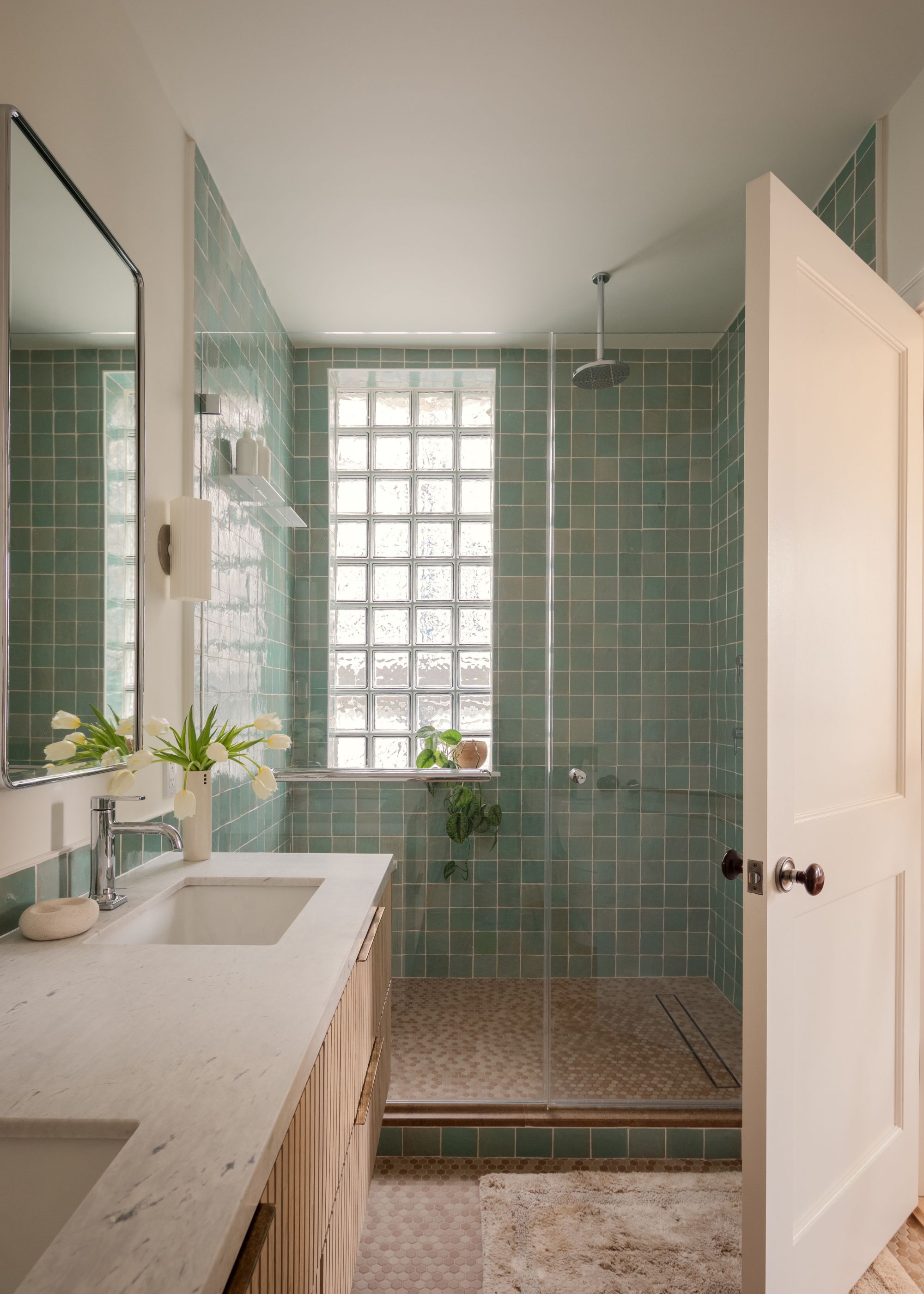 A modern, minimalist bathroom with sage green shower tiles, a glass brick window, double sinks with white stone counters, and a mirror on the wall.