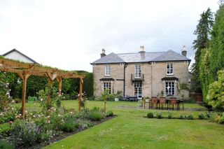 The house from the back garden shows a white block house with a balcony and grassy back garden with a pergola and a sitting area and a table with chairs with the garden barriers are hedges