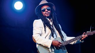 Julian Tayor wears dark sunglasses,a white blazer and a wide-brimmed hat as he plays his Custom Barncaster T-style onstage.