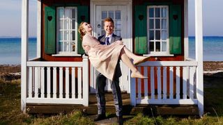 An attractive happy young husband carrying his beautiful new wife over threshold of cute, colourful beach hut for fun on the Danish island of Aero.