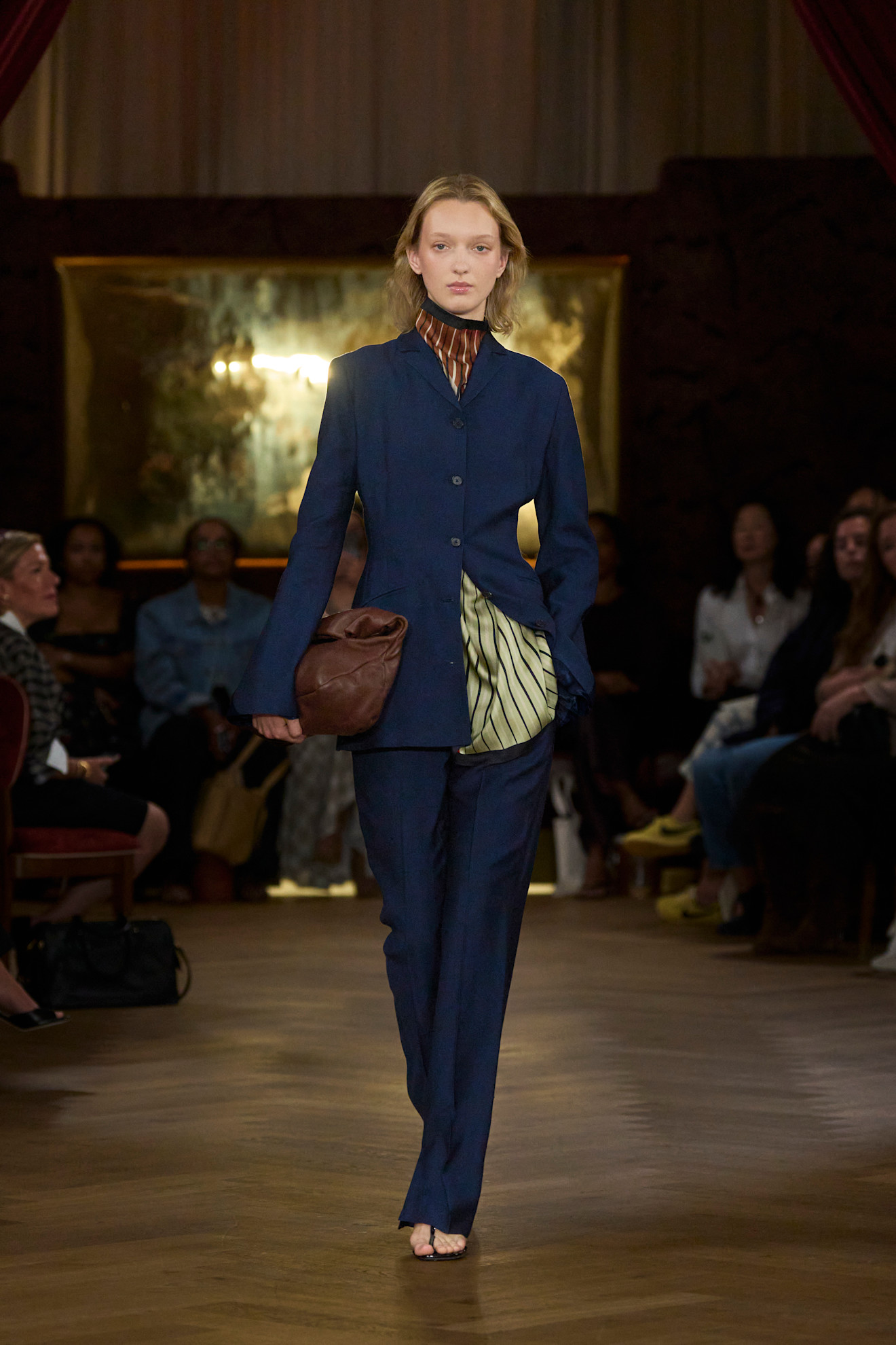 Layering staples for spring 2026 is shown in a photo from Kallmeyer's runway show, with a model walking while wearing a navy blazer styled with matching trousers styled with a two-tone scarf layered underneath, paired with black thong sandals and a brown hobo bag