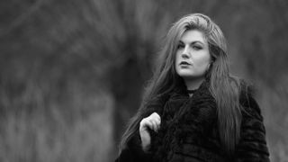 Black and white photograph of a woman, wearing glasses and a black coat, against a blurry outdoor background