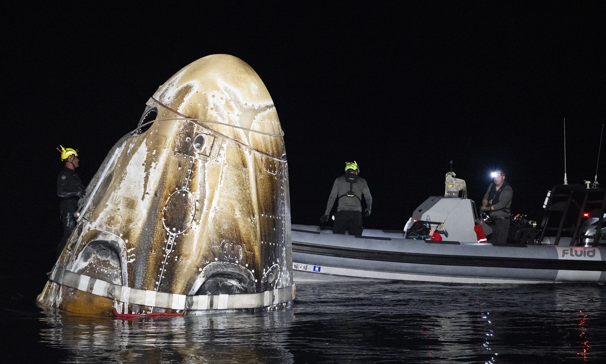 a charred cone-shaped spacecraft next to a small boat full of people, lit up in darkness