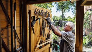 A man hangs tools on the door of a shed under the word 'garden'