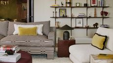 cozy modern living room with neutral walls, a brown check and striped sofa, brown side table and lamp, boucle chair with yellow velvet pillows and styled open shelving behind