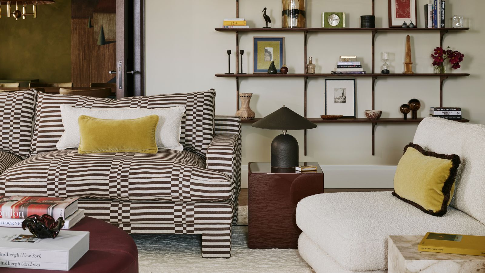 cozy modern living room with neutral walls, a brown check and striped sofa, brown side table and lamp, boucle chair with yellow velvet pillows and styled open shelving behind